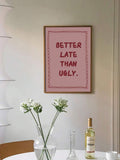 Framed BETTER LATE THAN UGLY pink typography print above dining table with flowers and wine