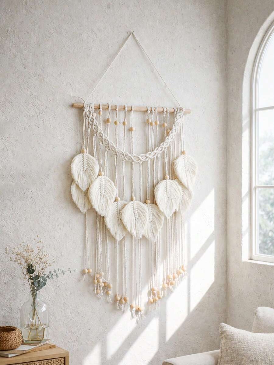 Cream macrame wall hanging with leaf tassels wooden beads and dowel on white wall beside hanging plant