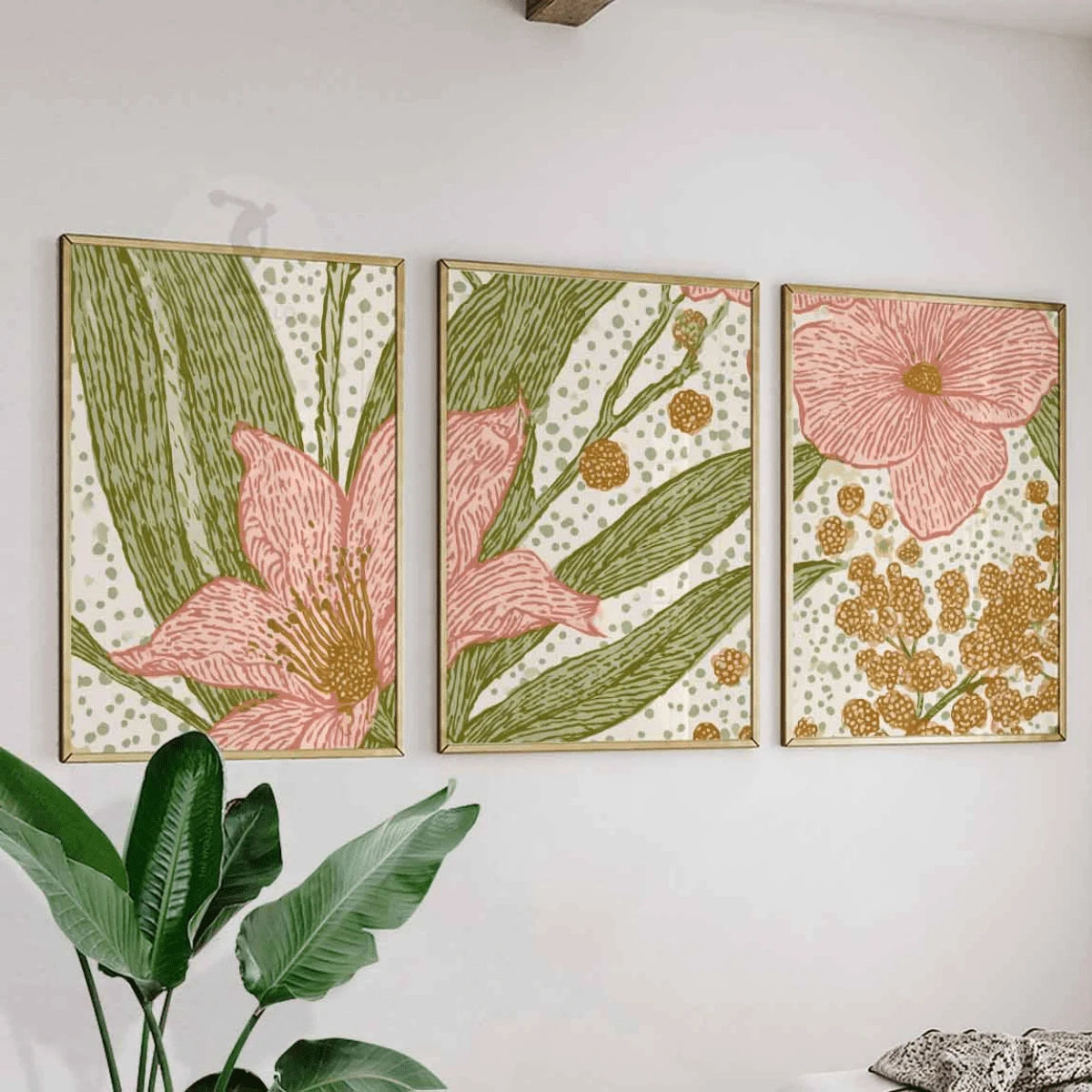 Three floral botanical panels in slim gold-toned frames on a white wall beside a broad-leaf houseplant