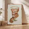 Framed luxury cocktail illustration displayed beside stacked books on a sunlit neutral wall