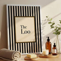 White framed The Loo print styled with towels soap candle amber bottles and greenery