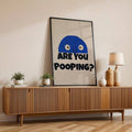Black framed humorous ARE YOU POOPING wall art on wooden sideboard with plants and lamp
