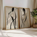 Two gold framed abstract figure prints leaning on floor beside plant and curtains
