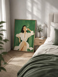 Framed woman drinking coffee artwork styled on bedroom floor beside nightstand and green bedding