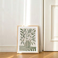 Framed olive vase botanical print leaning on wood floor near cream wall and door