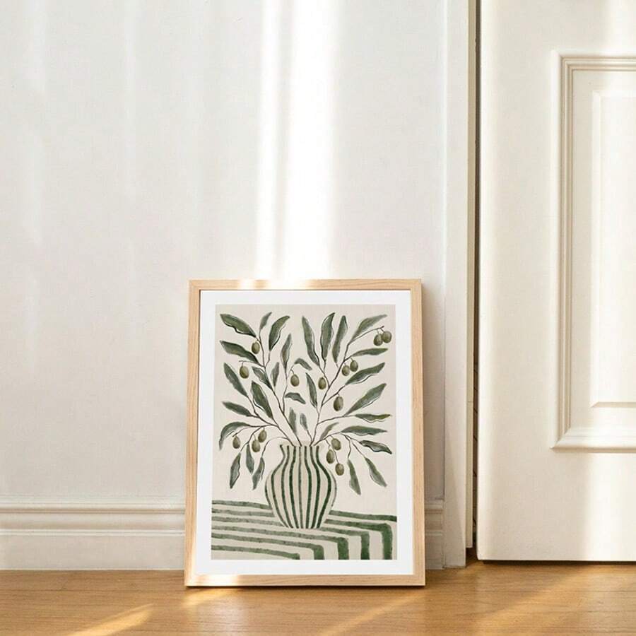 Framed olive vase botanical print leaning on wood floor near cream wall and door