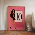 Gold framed pink fashion doll scoreboard poster leaning on floor beside books and chair