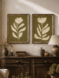 Two olive green botanical flower wall prints above rustic cabinet with books candle lamp vase and small pumpkin