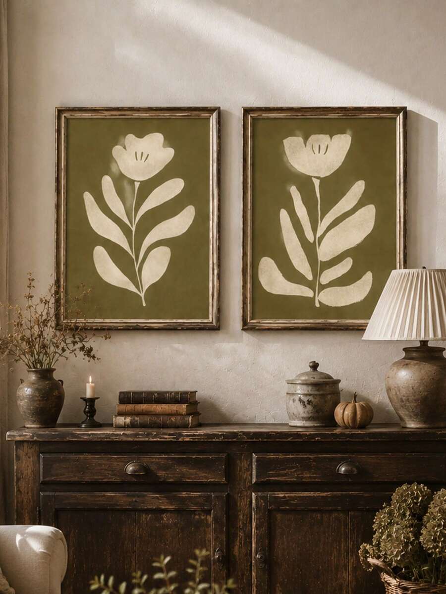 Two olive green botanical flower wall prints above rustic cabinet with books candle lamp vase and small pumpkin