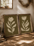 Two framed olive green botanical flower prints leaning on rustic wood table with pumpkin candle and dried vine decor