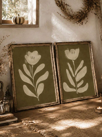 Two framed olive green botanical flower prints leaning on rustic wood table with pumpkin candle and dried vine decor