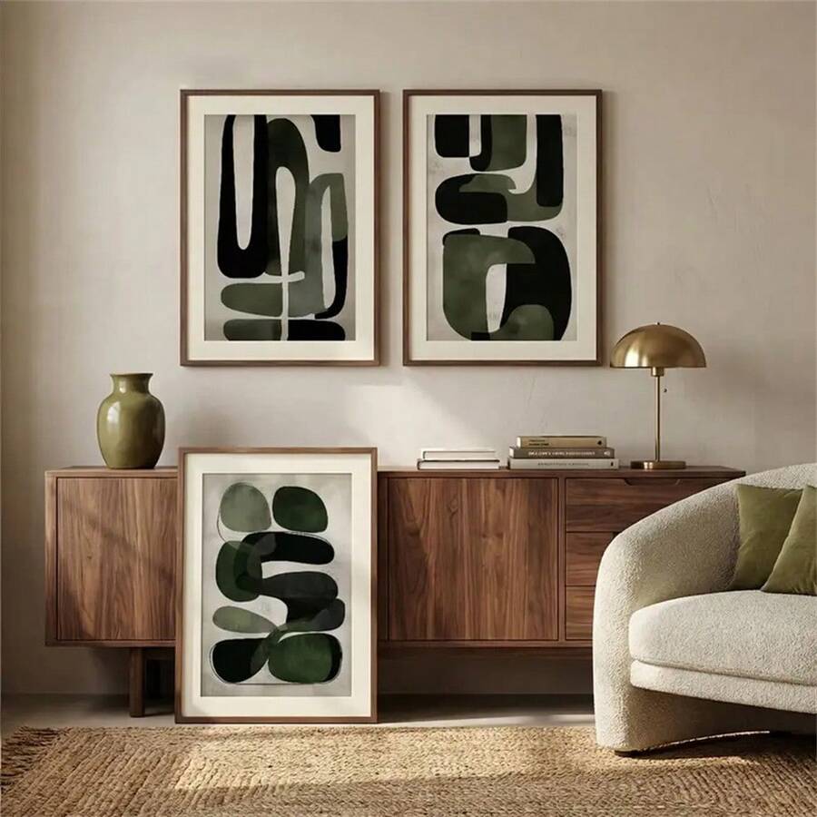 Two olive abstract prints hung with a third leaning beside a walnut credenza