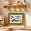 Framed folk-art fish print leaning on kitchen counter beneath patterned mugs