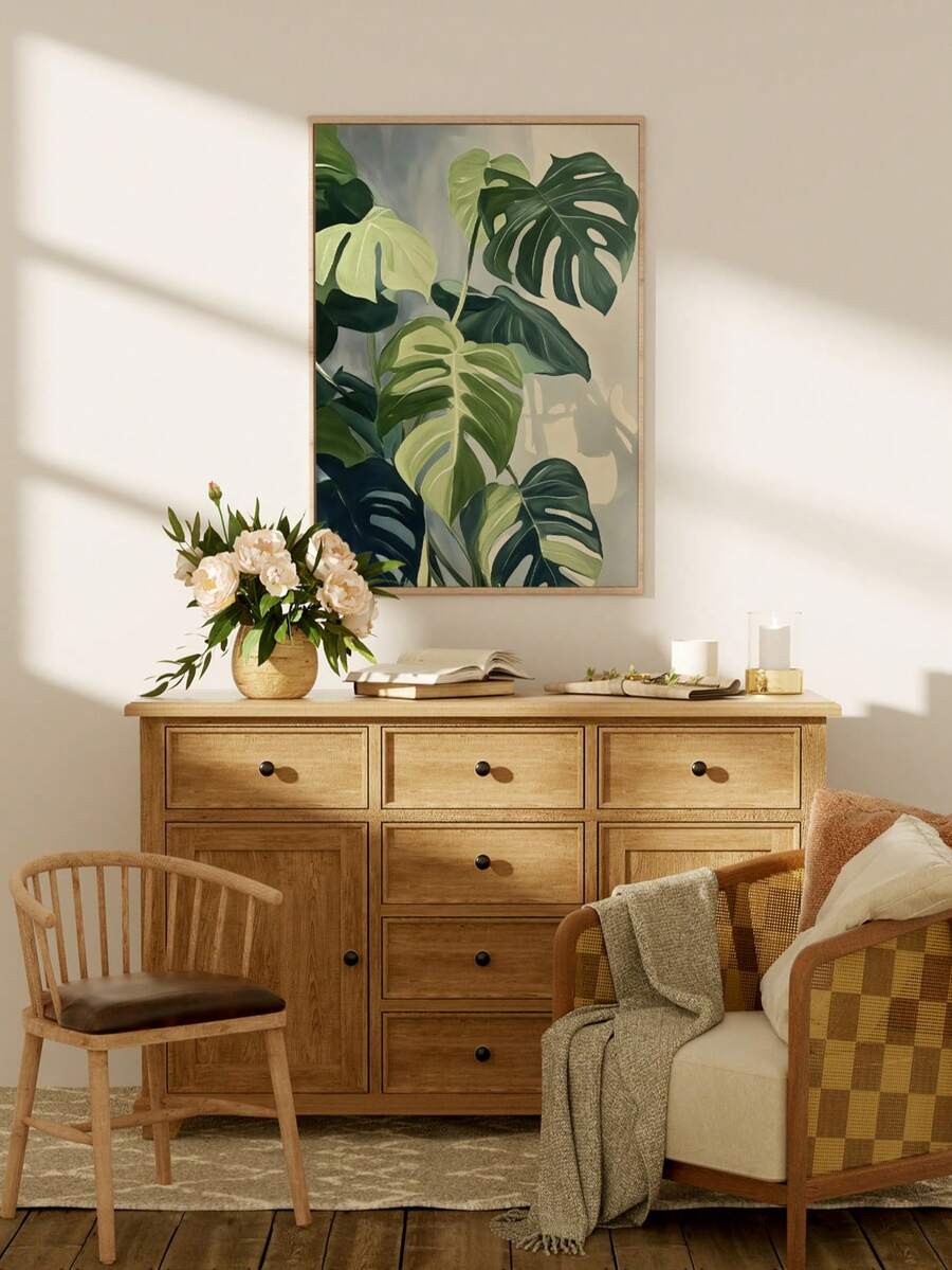 Framed monstera botanical wall art leaning against cream wall on pale wood floor in diagonal sunlight