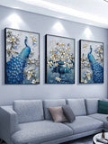 Three black framed blue peacock and white flower prints above gray sofa under spotlights