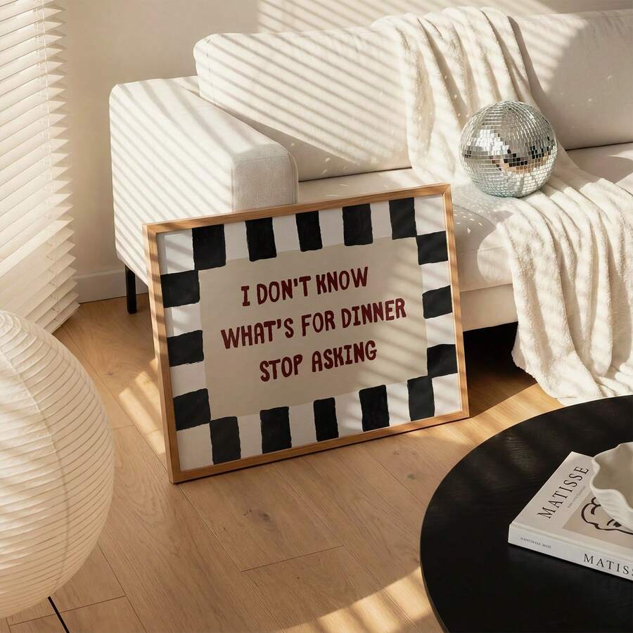 Humorous dinner quote print leaning beside cream sofa, white throw and silver disco ball