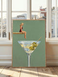 Blonde swimmer in burgundy suit perched above oversized dirty martini glass with green olives