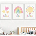 White-framed pastel wall art above nursery bed with Be brave hearts, Be you rainbow and Be happy sun