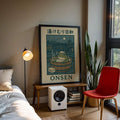 Black-framed onsen frog poster on a wood bench beside a lamp, books and red chair