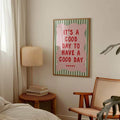 Good day quote wall art hanging beside a bed with a lamp, books, and cane chair
