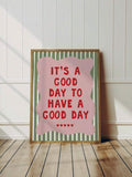 Framed good day quote print leaning against a white paneled wall on a wood floor