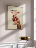 Gold-tone framed jeweled hand and pink coupe artwork on a sunlit paneled wall