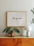 Framed love you more typography print above wood cabinet with plants