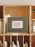 Cream blessing text panel on green gingham artwork displayed in a light wood bookshelf beside books