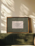 Dark green gingham scripture artwork in a brown frame displayed behind an olive green sofa in warm sunlight
