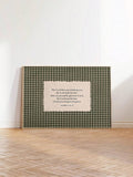 Green plaid blessing canvas artwork in a slim light frame leaning against a white wall on parquet flooring