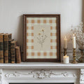 Dark wood framed daisy gingham artwork on a vintage mantel with books, candles, and dried flowers