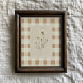 Dark wood framed daisy print with a beige gingham border on textured white linen