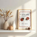 Light wood-framed ladybug wall art on a sunlit shelf with neutral vases and dried flowers