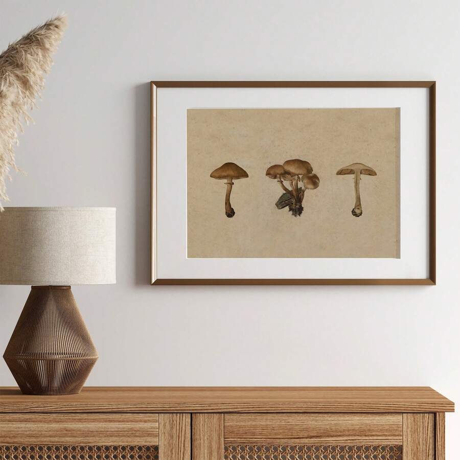 A framed art print featuring three brown mushrooms on a textured beige background, styled on a light gray wall above a wooden console table with a lamp.