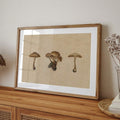 A framed art print featuring three illustrations of mushrooms on a textured beige background, styled on a wooden console table against a neutral wall.