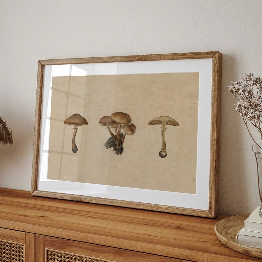 A framed art print featuring three illustrations of mushrooms on a textured beige background, styled on a wooden console table against a neutral wall.