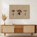 A framed mushroom art print with a beige background hangs on a white wall above a mid-century modern wooden sideboard with woven cane panels. A white ceramic vase with dried pampas grass and a stack of books sit on the cabinet.