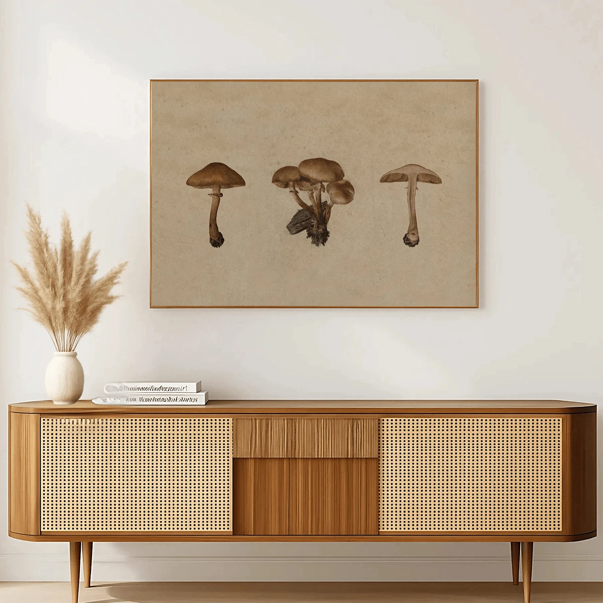 A framed mushroom art print with a beige background hangs on a white wall above a mid-century modern wooden sideboard with woven cane panels. A white ceramic vase with dried pampas grass and a stack of books sit on the cabinet.