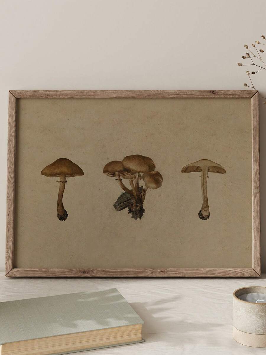 A framed botanical-style print showing three brown mushrooms on a warm beige background, enclosed in a thin gold-toned frame and photographed against a light neutral wall.