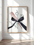 Watercolor scissors, pink comb and black bow artwork in a light wood frame on a bright wall beside a window