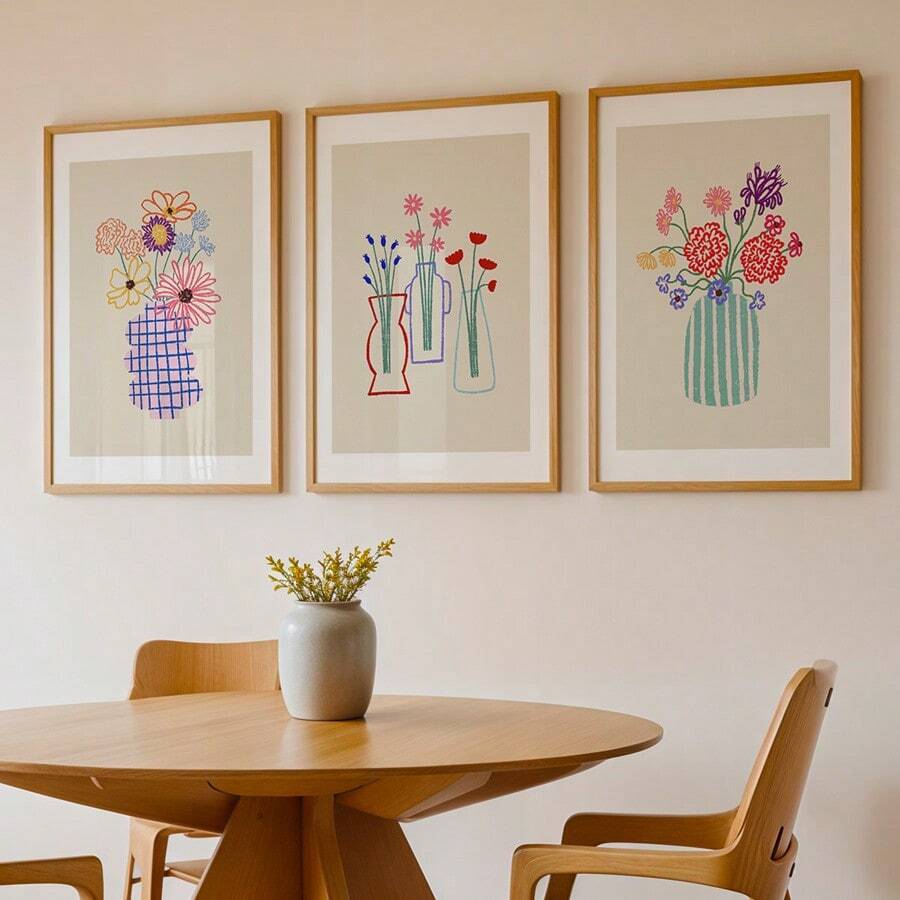 Three framed botanical vase prints above a dining table in a bright neutral room