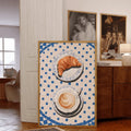 Framed croissant and coffee print displayed by a sunny window and potted plant
