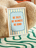 Framed Be Silly Be Honest Be Kind nursery canvas poster with blue stripes and orange typography beside a sofa