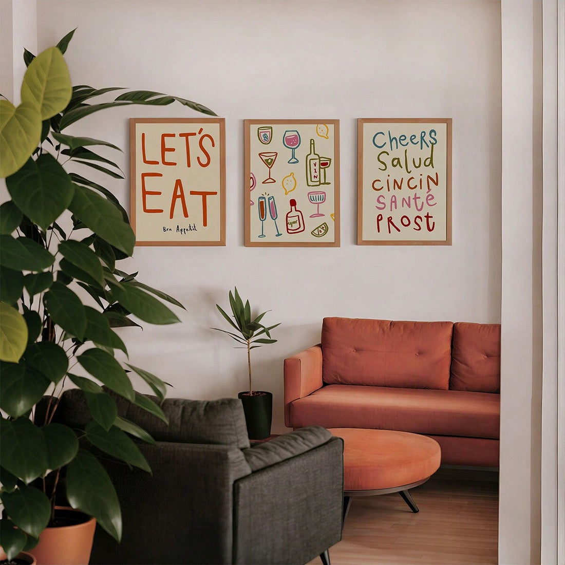 Three framed art prints with colorful text and illustrations hang on a white wall above a terracotta-colored sofa in a modern living room setting with a large green potted plant in the foreground.