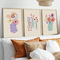 Three colorful hand-drawn floral vase canvases in pale wood frames above a white sofa