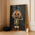 Orange pumpkin-headed skeleton artwork in a slim gold-toned display frame beside a black pumpkin and candle against a paneled wall