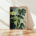 Framed monstera botanical wall art leaning against cream wall on pale wood floor in diagonal sunlight
