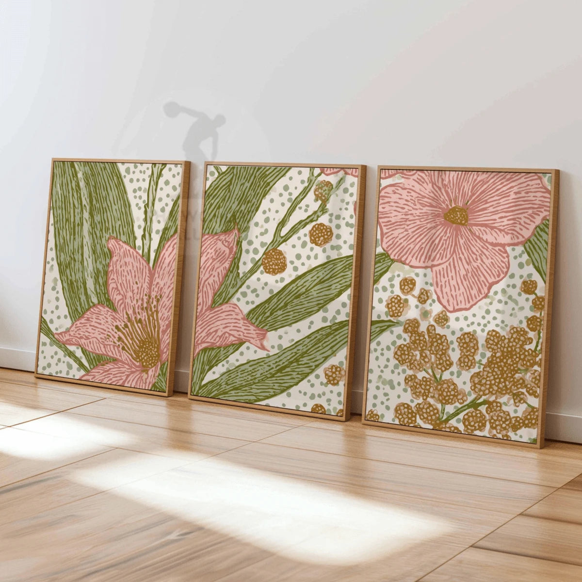 Three coordinated pink flower and mustard botanical prints in wood-toned frames leaning on a sunlit wooden floor