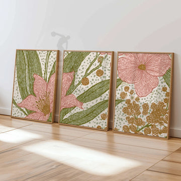Three coordinated pink flower and mustard botanical prints in wood-toned frames leaning on a sunlit wooden floor