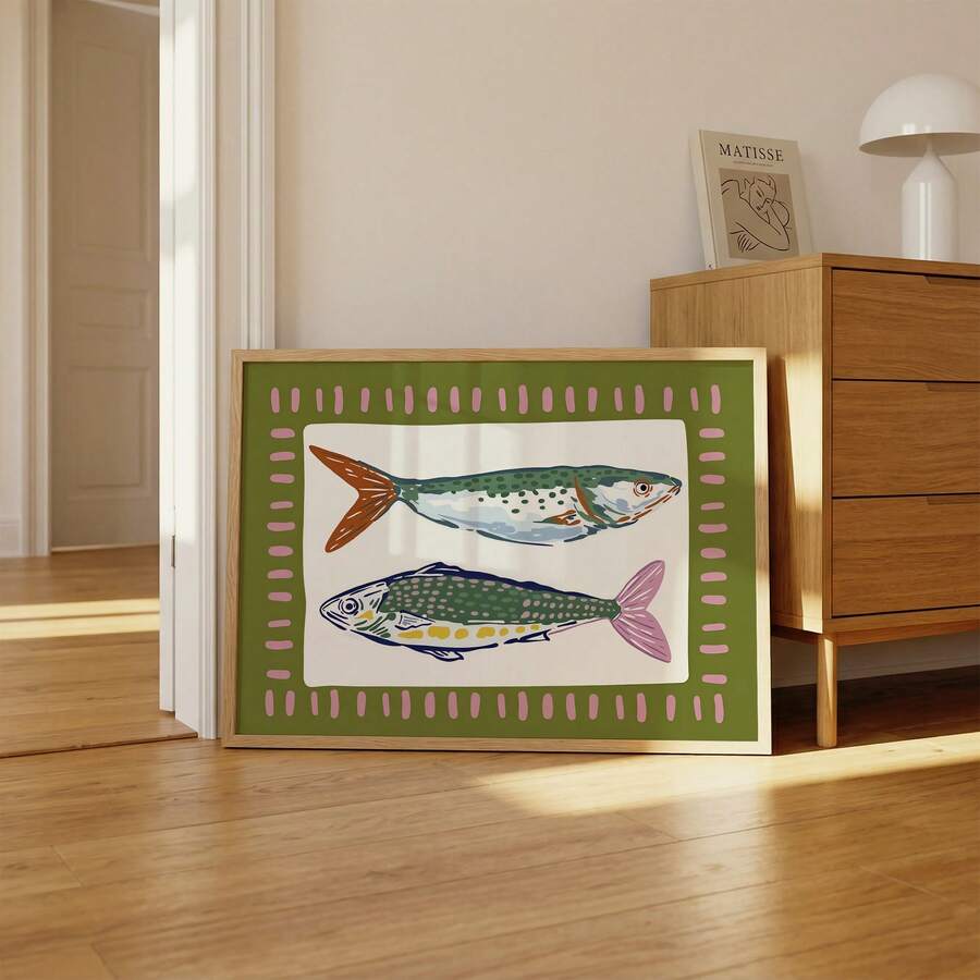 Framed two-fish coastal artwork leaning beside wood credenza in warm neutral room